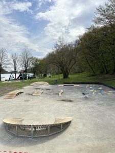 Ein Skatepark aus Beton mit Holzbänken, Rampen und bunten Motiven befindet sich im Freien in der Nähe von Bäumen, einem See und einem nahegelegenen Wanderweg unter einem teilweise bewölkten Himmel. Im Hintergrund ist ein Spielplatz zu sehen.