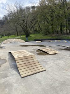 Ein Skatepark aus Beton mit mehreren, teilweise kaputten Holzrampen liegt unter einem bewölkten Himmel. Bäume mit spärlichem Laub und grünem Gras umgeben das Gelände und verleihen dem Hintergrund eine trailartige Atmosphäre.
