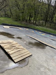 Auf einer Betonstrecke mit nassen Stellen, möglicherweise zum Mountainbiken oder Skaten, befinden sich Holzrampen und -bretter in der Nähe einer Rasenfläche und Bäumen im Hintergrund. Bunte Markierungen heben sich auf dem Boden ab.