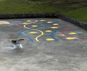 Eine Betonspielplatzfläche ist mit bunten Wellenlinien in Gelb, Blau, Grün und Rot versehen, die einer Spielbahn ähneln, die mehrere runde, gelbe Punkte miteinander verbindet. Am Rand steht eine Metallbank, hinter der niedrigen Betonkante ist Gras sichtbar.