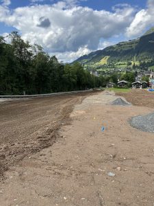 Eine Baustelle mit Erde und Schotter, kleine bunte Fähnchen markieren die Stellen – das perfekte Terrain für eine zukünftige Mountainbike-Strecke. Umgeben von Bäumen, grünen Hügeln in der Ferne, vereinzelten Häusern und einem leicht bewölkten Himmel prägen die Szenerie.