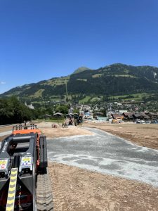 Eine Baustelle mit Maschinen im Vordergrund, unbefestigter Boden und ein teilweise asphaltierter Weg. Im Hintergrund liegt eine Stadt am Fuße grüner Berge unter einem klaren blauen Himmel – perfektes Terrain für Mountainbike-Enthusiasten.