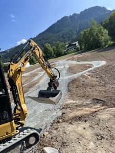 Ein gelber Bagger arbeitet auf einer Baustelle und verteilt Kies über einem zukünftigen Mountainbike-Trail, im Hintergrund sind grüne Bäume und Berge unter einem klaren blauen Himmel zu sehen.