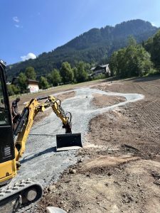 Ein gelber Bagger arbeitet auf einer Mountainbike-Strecke aus Erde und Schotter in einer ländlichen Gegend mit grünen Bäumen, Häusern und einem bewaldeten Berg unter einem blauen Himmel im Hintergrund.