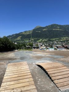 Ein Mountainbike-Trail mit zwei kleinen Holzrampen im Vordergrund überblickt eine Stadt, die von grünen Hügeln und Bergen unter einem klaren blauen Himmel umgeben ist.