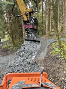 Eine Nahaufnahme eines Baggers, der aus seiner Schaufel nassen Beton auf einen Waldweg schüttet, im Hintergrund sind Bäume und Grünflächen zu sehen.