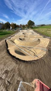 Ein im Bau befindlicher Pumptrack für Dirtbikes und Mountainbikes mit gewundenen, erhöhten Kurven und Hügeln aus gestampfter Erde, teilweise umgeben von Metallbarrieren, auf einer Wiese an einem klaren, sonnigen Tag.