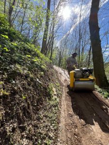 Eine Person bedient eine gelbe Verdichtungsmaschine auf einem schmalen Feldweg in einem Wald, wobei Sonnenlicht durch die hohen Bäume fällt und die üppige Vegetation den Hang bedeckt – perfekt, um die Bewegung mit dem Mountainbike zu fördern.