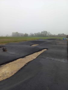 Ein asphaltierter Pumptrack mit erhabenen Kurven und unbefestigten Abschnitten schlängelt sich durch eine grasbewachsene, neblige Landschaft, ideal für Mountainbike-Enthusiasten, mit Bäumen und Feldern im Hintergrund.