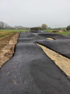 Ein neu asphaltierter Pumptrack mit sanften, welligen Kurven und Hügeln, perfekt für Mountainbike- oder MTB-Fans, umgeben von Gras und Erde an einem bewölkten Tag.