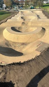 Ein unbefestigter BMX- oder Pumptrack im Bau mit Steilkurven, Hügeln und verschiedenen Steigungen – perfekt für Mountainbike- und Trail-Fans. Die Strecke ist von Gras umgeben, im Hintergrund sind Bauwerkzeuge und -materialien zu sehen.
