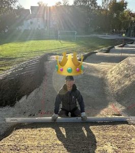 Eine Person hockt draußen auf einem sandigen Pfad und hält ein langes Planierwerkzeug in der Hand. Sonnenlicht fällt von hinten herein, und über ihrem Kopf prangt eine Cartoon-Krone. Im Hintergrund sind Gras und Bäume zu sehen – der wahre Geist der Mountainbike-Bewegung.