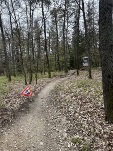 Ein Feldweg schlängelt sich durch einen Wald mit laubbedecktem Boden und hohen Bäumen. Links steht ein dreieckiges Warnschild, das auf Baustellen hinweist. Weiter unten arbeiten Freiwillige der Mountainbikebewegung an der Verbesserung des Mountainbike-Weges.