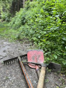 Drei Gartengeräte – ein roter Rechen, eine rote Schaufel und eine Spitzhacke – liegen auf einem Schotterweg neben dichtem grünem Laub und sind bereit, in einem Waldgebiet im Freien eine Mountainbike-Strecke anzulegen.
