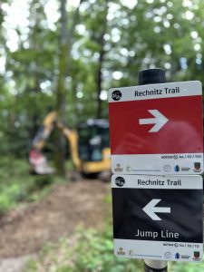 Eine Nahaufnahme zweier Wegweiser für den Rechnitz Trail mit nach rechts zeigenden Pfeilen, darunter einer mit der Aufschrift „Jump Line“ – perfekt für jeden MTB-Fan. Im Waldhintergrund ist ein unscharfer Bagger zu sehen, der auf aktive Gleisarbeiten hinweist.