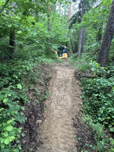 Ein schmaler Feldweg schlängelt sich durch dichten grünen Wald bergauf und führt zu einem kleinen gelben Bagger, der im Hintergrund teilweise zwischen den Bäumen zu sehen ist – ideales Terrain für Mountainbike-Abenteuer.
