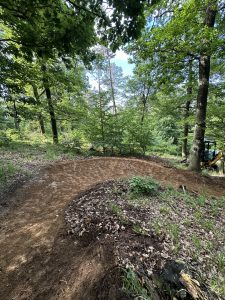 Eine unbefestigte Mountainbike-Strecke schlängelt sich durch einen Wald mit hohen, grünen Bäumen. Frisch gerodete Erde markiert den Weg, und zwischen den Bäumen auf der rechten Seite ist teilweise ein kleiner Bagger zu erkennen.
