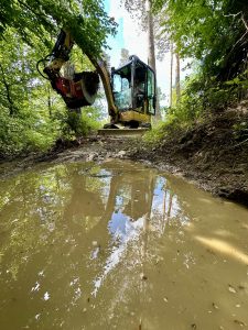 Ein gelber Bagger steht auf einem schlammigen Waldweg, sein Spiegelbild ist in einer großen Pfütze zu sehen – ein beliebtes Gebiet für Mountainbike-Fans, die die umliegenden grünen Bäume und das Unterholz erkunden.