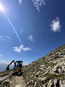 Ein kleiner Bagger steht auf einem felsigen Berghang neben einem Mountainbike-Trail unter einem strahlend blauen Himmel mit vereinzelten Wolken und Sonnenlicht, das aus der oberen linken Ecke strömt.