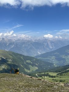 Ein gelber Bagger steht auf einem grasbewachsenen Hügel in der Nähe eines gewundenen Pfades und überblickt ein üppig grünes Tal mit Bergen in der Ferne unter einem teilweise bewölkten blauen Himmel.