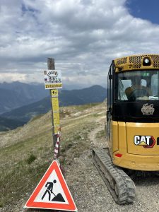 Ein gelber CAT-Bagger steht auf einem felsigen Bergpfad neben einem Bike-Zirbentrail-Schild und einem Warnschild für Straßenarbeiten und weist auf die Mountainbike-Bewegung in der Gegend hin, mit Bergen und einem bewölkten Himmel im Hintergrund.