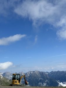 Ein gelber Bagger steht auf felsigem Boden in der Nähe einer schroffen Mountainbike-Strecke, im Hintergrund sind schneebedeckte Berge und ein blauer Himmel mit vereinzelten Wolken zu sehen.