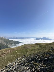 Eine malerische Aussicht von einem felsigen Hügel mit Blick auf einen grasbewachsenen Berghang und einen gewundenen Pfad, mit einer Wolkenschicht, die das Tal darunter bedeckt, und in der Ferne Berggipfel unter einem klaren blauen Himmel.
