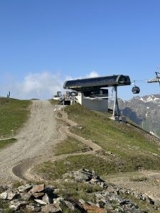 Auf einem grasbewachsenen Berggipfel befindet sich eine Seilbahnstation, zu der ein gewundener Pfad hinaufführt. Auf der rechten Seite hängt eine Seilbahn, und in der Ferne sind unter einem teilweise klaren Himmel felsige Gipfel zu sehen.