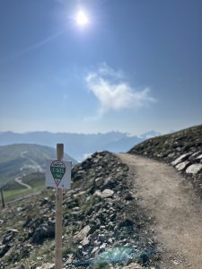 Ein unbefestigter Wanderweg schlängelt sich bei strahlendem Sonnenschein und klarem Himmel durch felsiges Gelände bergauf. Ein Holzpfosten mit rot-grüner Wegmarkierung säumt den Weg – ideal für Wanderer und Mountainbiker, mit den Bergen in der Ferne im Hintergrund.