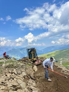 Eine Person bearbeitet mit einer Schaufel eine steinige Mountainbike-Strecke, während in der Nähe ein kleiner Bagger arbeitet. Im Hintergrund sitzt eine weitere Person auf Felsen. Der blaue Himmel ist von gewaltigen Bergen und Wolken geprägt.