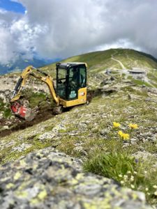 Ein gelber Bagger arbeitet unter einem bewölkten Himmel an einem felsigen, grasbewachsenen Berghang. Im Vordergrund sind gelbe Wildblumen zu sehen, und im Hintergrund führt ein gewundener Mountainbike-Pfad zu einem Gebäude.