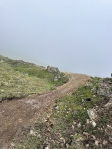 Ein gewundener unbefestigter Mountainbike-Pfad führt durch grasbewachsenes, felsiges Gelände an einem Hang und verschwindet im dichten Nebel, der den Hintergrund und den Himmel verdeckt.