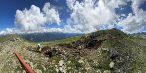 Bagger arbeiten an einem felsigen Hang unter leicht bewölktem Himmel. Im Hintergrund sind grüne Berge und Täler zu sehen. Ein großes orangefarbenes Rohr verläuft über den Boden und markiert möglicherweise die Route eines zukünftigen Mountainbike-Trails oder -Pfades.