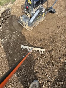 Ein Metallrechen verteilt Erde und Steine auf dem Boden und formt so einen zukünftigen Mountainbike-Trail, während eine Rüttelplatte daneben steht. Am unteren Bildrand ist ein Teil eines Stiefels zu sehen.