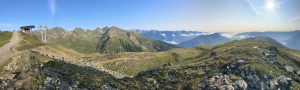 Panoramablick auf eine Berglandschaft mit felsigen Hängen, Grasflächen und einer Seilbahnstation auf der linken Seite. Ein gewundener Pfad schlängelt sich durch die Szene, während sich unter einem klaren blauen Himmel zerklüftete Gipfel am Horizont erstrecken.