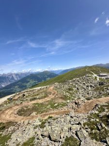 Ein gewundener Feldweg führt durch eine felsige Berglandschaft unter einem klaren blauen Himmel, im Hintergrund sind grüne Hügel und schneebedeckte Gipfel zu sehen – perfekt für ein Mountainbike-Abenteuer.