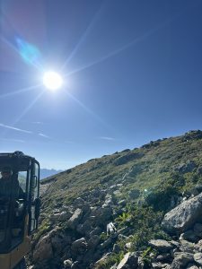 Eine strahlende Sonne scheint vom klaren blauen Himmel über einem felsigen, grasbewachsenen Hang – die ideale Kulisse für jedes Mountainbike-Abenteuer. In der unteren linken Bildecke sind Teile eines Baufahrzeugs und sein Fahrer zu sehen.