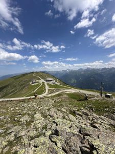 Eine kurvenreiche Mountainbike-Strecke schlängelt sich unter einem strahlend blauen Himmel mit vereinzelten Wolken einen grasbewachsenen Hügel hinauf, umgeben von felsigem Gelände und fernen Berggipfeln.