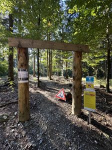 Ein unbefestigter Waldweg führt unter einem Holztor aus Baumstämmen hindurch. Sonnenlicht fällt durch die grünen Bäume. Warnschilder, Informationstafeln und ein rotes Dreieck kennzeichnen den Wegeingang für Mountainbiker.