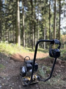 Eine kompakte Rüttelplatte steht auf einem Feldweg in einem sonnenbeschienenen Wald. Gehörschutzkapseln hängen am Griff, und hohe Bäume mit grünem Laub prägen den Hintergrund – perfekt für Ihr nächstes Mountainbikemovement-Abenteuer.