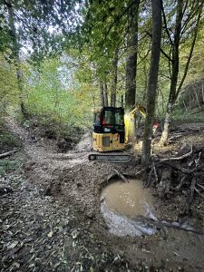Ein kleiner gelber Bagger gräbt in der Nähe einer schlammigen Pfütze in einem Waldgebiet, umgeben von Bäumen mit grünen Blättern und unebenem, mit Schlamm und Laub bedecktem Gelände entlang eines Mountainbike-Trails.