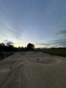Ein weites, sandiges Feld unter einem sich verdunkelnden Abendhimmel mit flachen Hügeln und Senken, perfekt für Mountainbike-Fahrten. Vereinzelte Bäume säumen den Horizont, und rechts erstrecken sich Grasfelder – ideales Terrain für jedes Mountainbike-Abenteuer.
