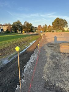 Eine Baustelle mit Schotter, Schlammflächen und einer roten Leitlinie deutet auf eine zukünftige Mountainbike-Strecke hin. Pfosten mit Tennisbällen markieren die Grenze, während im Hintergrund grünes Gras und Bäume unter einem strahlend blauen Himmel hervorstechen.