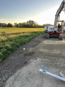 Eine Baustelle bei Sonnenuntergang zeigt einen mit Schnur und Pfählen markierten Schotterweg neben einer Wiese. Ein Bagger und ein weißer Lastwagen parken in der Nähe. Im Vordergrund sind Werkzeuge für die Fortbewegung eines Mountainbikes zu sehen.