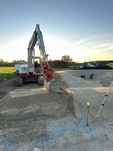 Eine Baustelle bei Sonnenuntergang zeigt einen Bagger, der Kies auf einer unbefestigten MTB-Strecke verteilt, mit Baumaschinen im Hintergrund, markierten Grenzen und offenen Feldern rund um das Gebiet – die Bühne für Mountainbikemovement-Abenteuer.