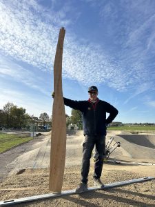 Eine Person mit Sonnenbrille und Mütze steht lächelnd auf einem Sandweg und hält ein hohes, gebogenes Holzbrett aufrecht. Der Himmel ist teilweise bewölkt, im Hintergrund sind Werkzeuge und Grünflächen zu sehen.