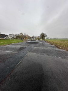 Ein frisch asphaltierter, leerer BMX-Pumptrack mit sanften Hügeln und Kurven an einem bewölkten, trüben Tag, umgeben von Gras, ein paar Häusern und Bäumen in der Ferne – perfekt für jeden Mountainbike- oder Mountainbikebewegungs-Enthusiasten.