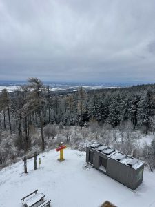Eine verschneite Landschaft mit Kiefern, einem kleinen grauen Gebäude, Picknicktischen und einem rot-gelben Wegweiser markiert den Beginn eines Mountainbike-Trails unter einem bewölkten Himmel mit fernen Hügeln im Hintergrund.