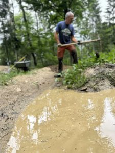 Eine Person schaufelt auf einer schlammigen Mountainbike-Strecke neben einer großen Pfütze, umgeben von grünen Bäumen und Pflanzen. Im Hintergrund ist eine Schubkarre zu sehen, der Fokus liegt auf der Pfütze im Vordergrund.