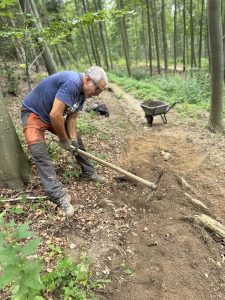 Ein Mann in Arbeitshandschuhen und Outdoor-Kleidung gräbt mit einer Hacke eine Mountainbike-Strecke im Wald. Hinter ihm steht eine Schubkarre auf dem Weg zwischen hohen Bäumen und viel Grün und zeigt damit seine Beteiligung an der Mountainbike-Bewegung.